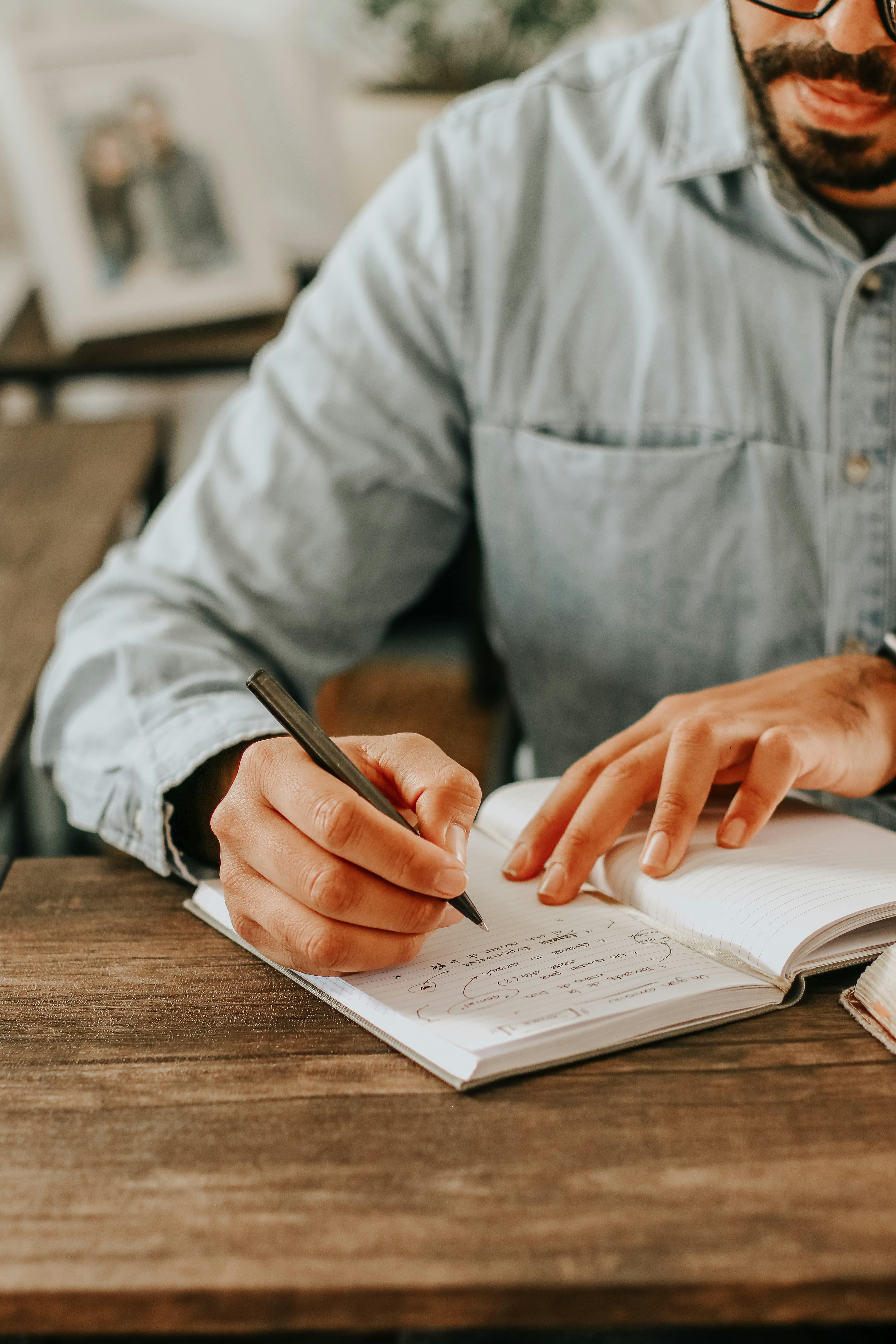 A person in a denim shirt writing in a lined journal at a wooden table