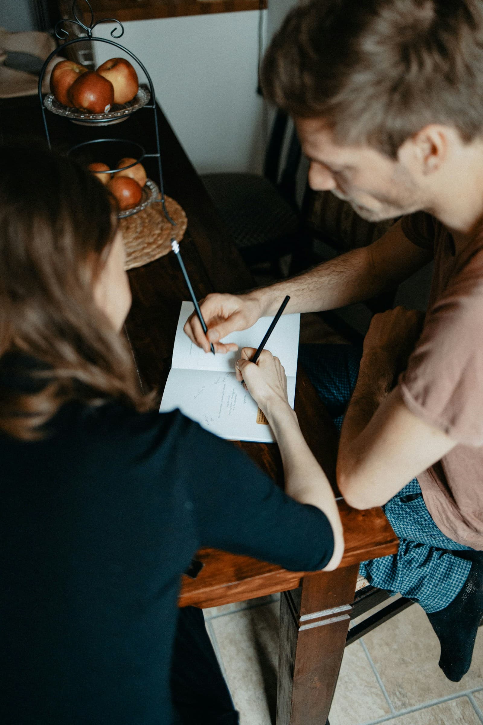 A couple sitting at a table together, each writing in a shared notebook